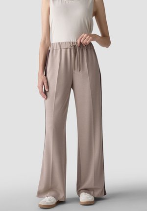 Model trägt beige weite Hosen mit schwarzen Seitenstreifen, elastischem Bund mit Kordelzug, weiße Sneaker und ein ärmelloses weißes Top.