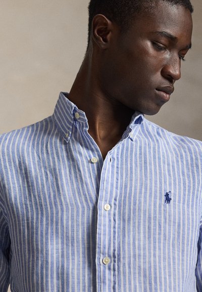 Άνδρας που φοράει ένα πουκάμισο με κάθετες ρίγες σε ανοιχτό μπλε και λευκό, με μικρό κεντημένο λογότυπο αλόγου στο στήθος, κοιτάζοντας προς τα κάτω.
