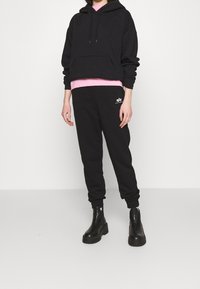 Svart huva tröja i lager över en rosa skjorta, kombinerad med svarta avsmalnande mjukisbyxor. Svarta ankelstövlar fulländar outfiten.