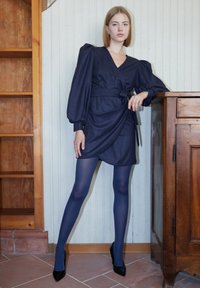 Abito avvolgente blu scuro con maniche a sbuffo e dettaglio sulla cintura; abbinato a collant blu scuro e décolleté neri a punta. Sfondo in legno e scaffale visibili.