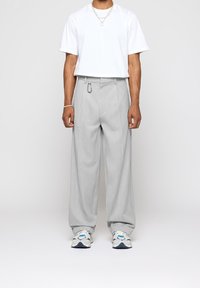 Pantalon gris clair à jambes larges avec une texture lisse, devant plissé et anneau détachable. Associé à un t-shirt blanc à manches courtes et des chaussures.