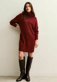 Robe pull col montant bordeaux avec manches longues et ourlet côtelé, associée à de hautes bottes en cuir marron pour un look superposé.