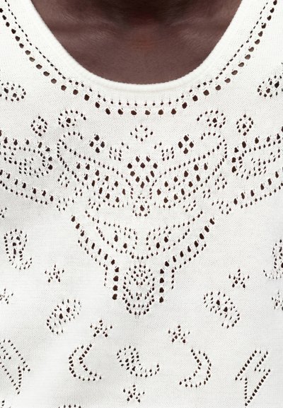 Κοντινό σε σκούρο δέρμα που φοράει λευκό πλεκτό τοπ με περίπλοκο σχέδιο από κεντήματα και στρογγυλή λαιμόκοψη.