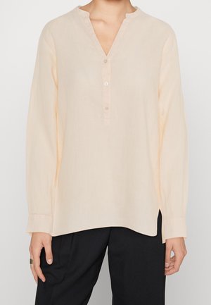 Mujer con blusa de manga larga color beige claro con cuello en V y tres botones, combinada con pantalones negros sobre un fondo liso.