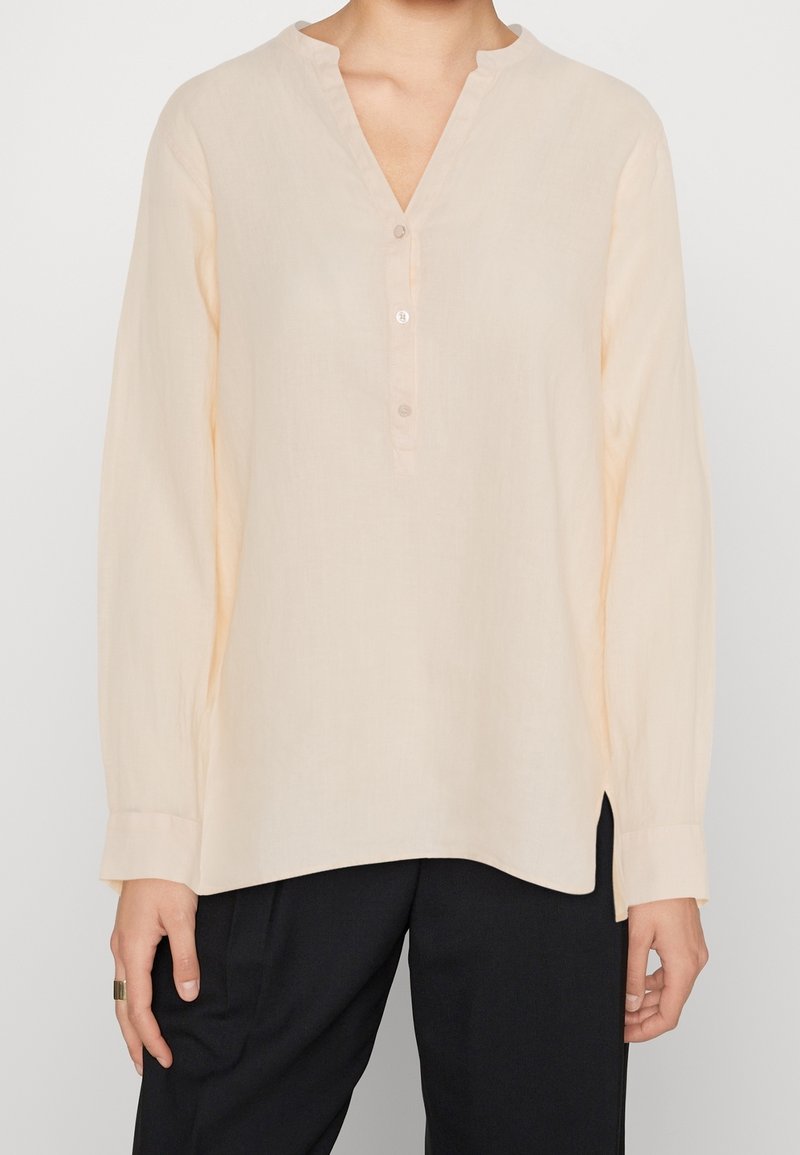 Femme portant un chemisier beige clair à manches longues avec un col en V et trois boutons, associé à un pantalon noir sur un fond uni.