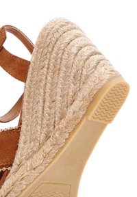 Wedge sandaal met een beige geweven jute zool, bruine suède banden en subtiele gouden kralenaccenten langs de band. Textuur rubberen loopzool.