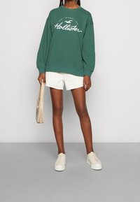 Vihreä huppari, jossa valkoinen kirjainlogo, yhdistetty valkoisiin shortseihin ja beigen värisiin lenkkareihin. Malli pitää käsissään vaaleanväristä olkalaukkua.