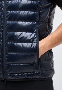 Χέρι που φοράει μαύρο γυαλιστερό γιλέκο με ραμμένα σχέδια, με το χέρι μέσα στην τσέπη με φερμουάρ στο πλάι, εμφανίζεται πάνω σε ανοιχτό φόντο.