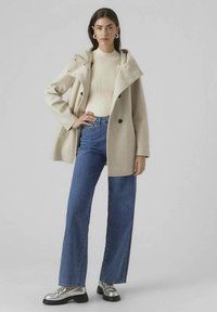 Bēšas vilnas maisījuma mētelis ar kapuci, pogu aizdari un platām piedurknēm, valkāts pār krēmkrāsas ribotu džemperi un ziliem augsta vidukļa džinsiem. Sudraba loaferi.