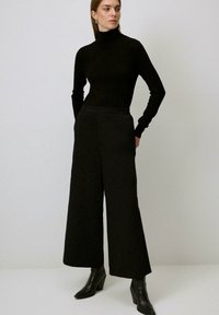 Femme debout les mains dans les poches, portant un col roulé noir, un pantalon large noir et des bottines à talons noires sur fond uni.