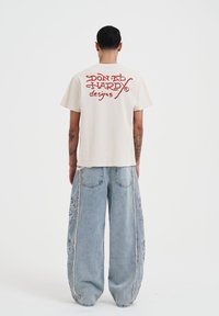 Krémové bavlněné tričko s červeným textem "Don Ed Hardy designs" na zádech; spárované s volnými světle modrými džíny se vzorovanými bočními panely.