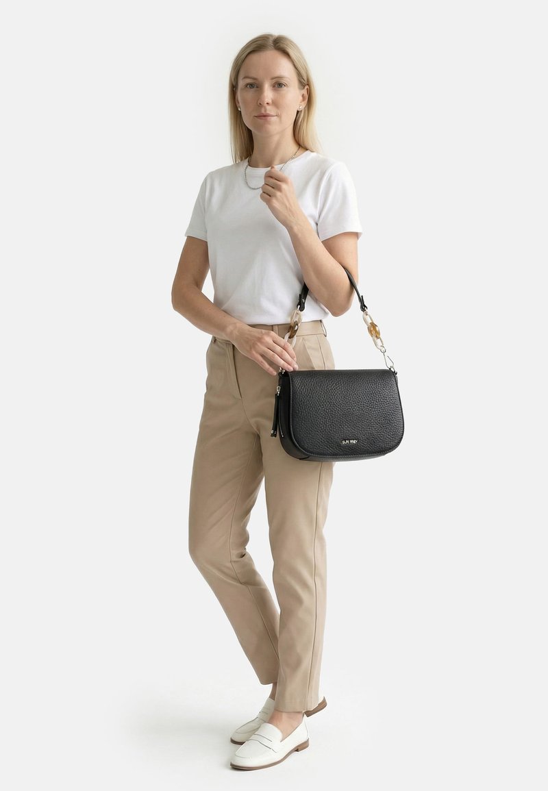 Frau trägt weißes T-Shirt und beigen Hosen, hält eine schwarze Lederhandtasche mit Schildpattkettendetails.