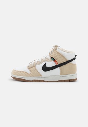 Basket montante blanche et beige avec le swoosh Nike noir, lacets blancs et semelle en caoutchouc marron sur fond blanc.