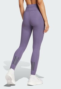 Leggings en tissu violet avec taille haute, dotés de trois rayures verticales noires sur le bas de la jambe, accompagnés de baskets blanches.