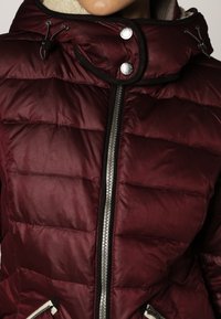 Veste matelassée bordeaux avec fermeture éclair à l'avant, accents de boutons et col haut. Présente une texture matelassée horizontale et des poches latérales.
