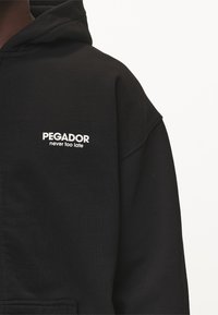 Felpa nera con cappuccio e vestibilità comoda. Presenta un logo testuale bianco "PEGADOR never too late" sulla parte frontale. Texture morbida, design casual.