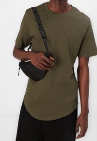 Camiseta de algodón verde oliva con dobladillo redondeado, combinada con un pequeño bolso cruzado negro que presenta una correa con logo y cierre de cremallera.