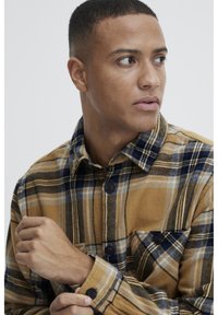 Brown and navy plaid flannel shirt featuring a button-down collar, chest pocket, and black buttons. Soft texture with visible fabric patterns.