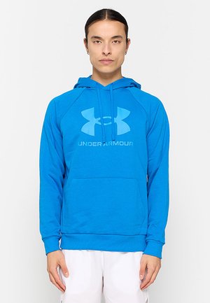 Personne portant un sweat à capuche Under Armour bleu vif avec un grand logo et texte, associé à un pantalon blanc, debout sur un fond blanc uni.