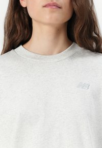 Sweatshirt gris clair avec un col ras du cou, arborant un petit logo New Balance brodé dans une teinte légèrement plus foncée près de la poitrine. Texture douce.