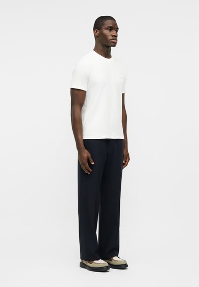 Άνδρας που στέκεται φορώντας λευκό κοντομάνικο μπλουζάκι, μαύρο φαρδύ παντελόνι και μπεζ μοκασίνια, σε απλό ανοιχτό φόντο.