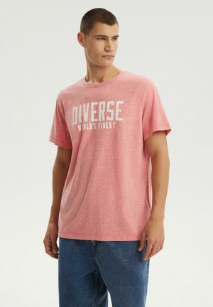 Młody mężczyzna ubrany w różowy t-shirt z napisem "DIVERSE WORLD'S FINEST" i niebieskie dżinsy, patrzący w bok na jednolitym tle.