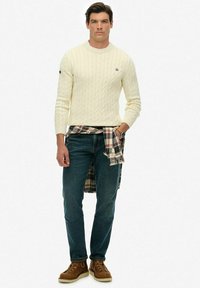 Suéter de punto trenzado crema, jeans oscuros, camisa de cuadros atada a la cintura y botas marrones con cordones. Diseño minimalista con tejido texturizado.