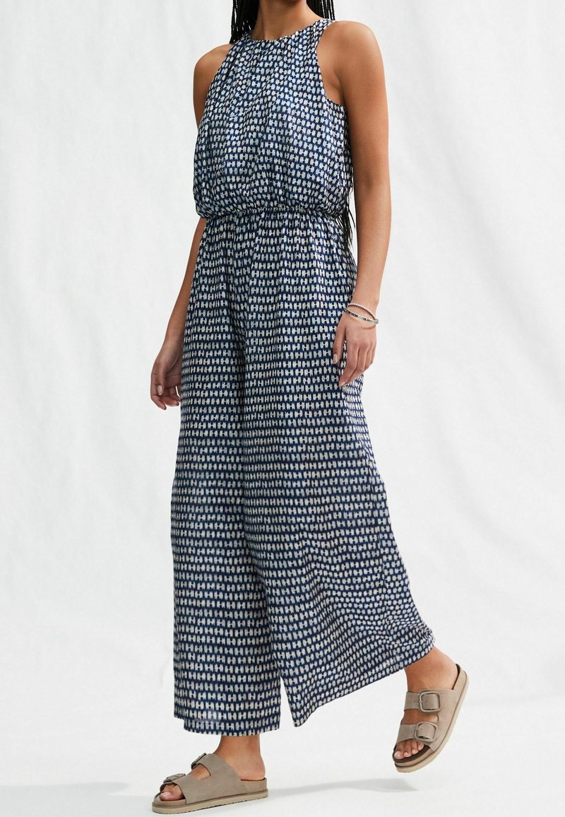 Femme portant une combinaison sans manches à jambes larges avec un motif bleu marine et blanc, des sandales beiges à boucle et des bracelets à un poignet.