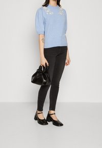 Helles, blaues Strickoberteil mit besticktem Blumenmuster, kombiniert mit schwarzen, figurbetonten Jeans und einer kleinen schwarzen Handtasche. Das Model trägt schwarze High Heels.