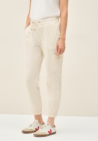 Beige elastické jogger kalhoty se dvěma p�ředními kapsami a manžetami na nohavicích, spárované s bílými teniskami s červenými akcenty a béžovými detaily.