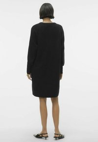 Abito in maglia oversize nero con maniche lunghe, orlo a coste e una consistenza morbida. Abbinato a scarpe nere con tacco basso. Vista da dietro.
