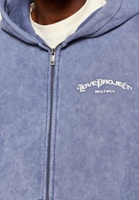 Svetle modrá mikina na zip vyrobená z měkké látky. Obsahuje přední zip, nastavovací kapuci a bílý potisk s textem "LOVE PROJECT" a "MULTIPLY."