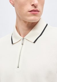 Άνδρας που φοράει ένα κρεμ πόλο με φερμουάρ μπροστά και μαύρο περίγραμμα στο γιακά, δείχνοντας το ντεκολτέ και μέρος του στήθους.