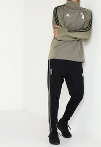 Sportoberteil von Adidas in olivgrün mit Stehkragen, schwarzem und weißem Logo, und schwarzen Jogginghosen mit seitlichen Streifen und Logodetails.