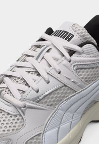 Λευκό και γκρι αθλητικό παπούτσι Puma με πάνελ διχτυού, λευκά κορδόνια και μαύρη επένδυση στο εσωτερικό, εμφανιζόμενο σε κοντινή λοξή όψη.