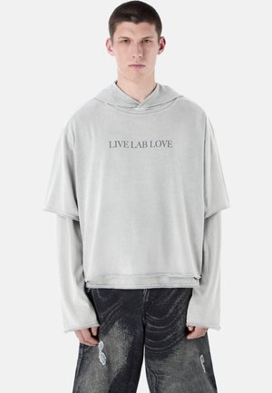 Młody mężczyzna w jasnoszarej, oversize'owej bluzie z napisem "LIVE LAB LOVE" i czarnych, przetartych dżinsach na jednolitym tle.