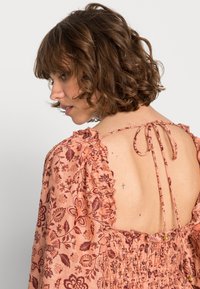 Vestido de padrão floral em pêssego com mangas franzidas e laço nas costas. Apresenta detalhe de franzido e acabamentos em metal dourado.