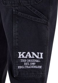 Czarne spodnie dżinsowe z kieszenią typu cargo, na której znajduje się biały haftowany napis: "KANI THE ORIGINAL EST. 1989 REG. TRADEMARK." Napis jest wyśrodkowany.