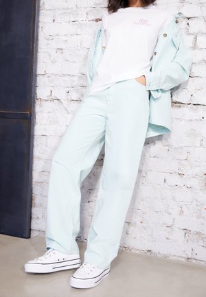 Osoba ubrana w jasnoniebieskie spodnie i kurtkę na białej koszulce z nadrukiem oraz białe sneakersy na platformie, stoi na tle białej ceglanej ściany.