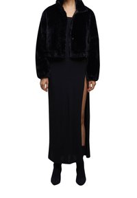 Kvinde iført en sort faux fur cropped jakke over en sort maxi kjole med en høj slids og sorte ankelstøvler.