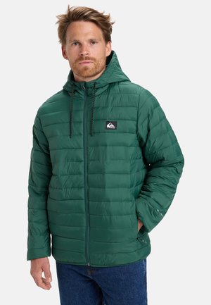 Homme portant une veste à capuche matelassée verte avec fermeture éclair à l'avant et poches latérales, se tenant debout contre un fond clair uni.
