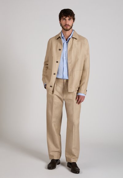 Homme debout portant un costume beige avec une veste et un pantalon large, une chemise bleu clair et des chaussures marron foncé sur un fond uni.