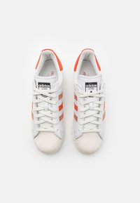 Sneakers bianchi Adidas con dettagli arancioni, dotati di puntale testurizzato, tre strisce e lacci piatti. Design classico con colletto imbottito.