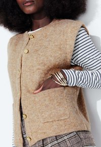 Gilet en laine beige avec une finition texturée, fermeture à boutons sur le devant et poches latérales. Porté sur une chemise à rayures à manches longues.
