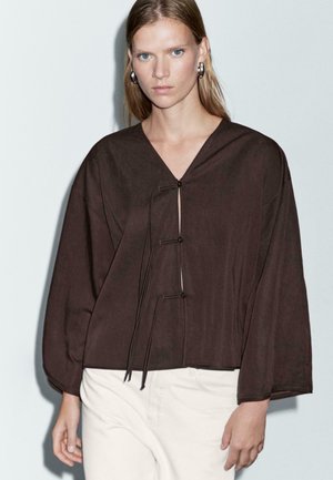 Femme blonde portant un manteau en cuir noir surdimensionné avec des manches larges et un pantalon clair, se tenant devant un fond clair.