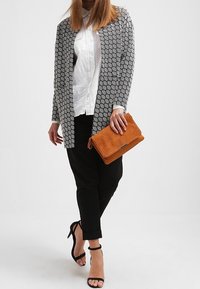 Sort og hvid mønstret åben cardigan, hvid skjorte, sorte bukser, holder en orange tekstureret clutch og bærer sorte remsandaler med hæl.