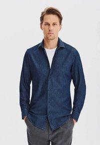 Granatowa koszula dżinsowa z zapięciem na guziki, długimi rękawami i zaokrąglonym dołem. Nosi się ją na białej koszulce z okrągłym dekoltem.