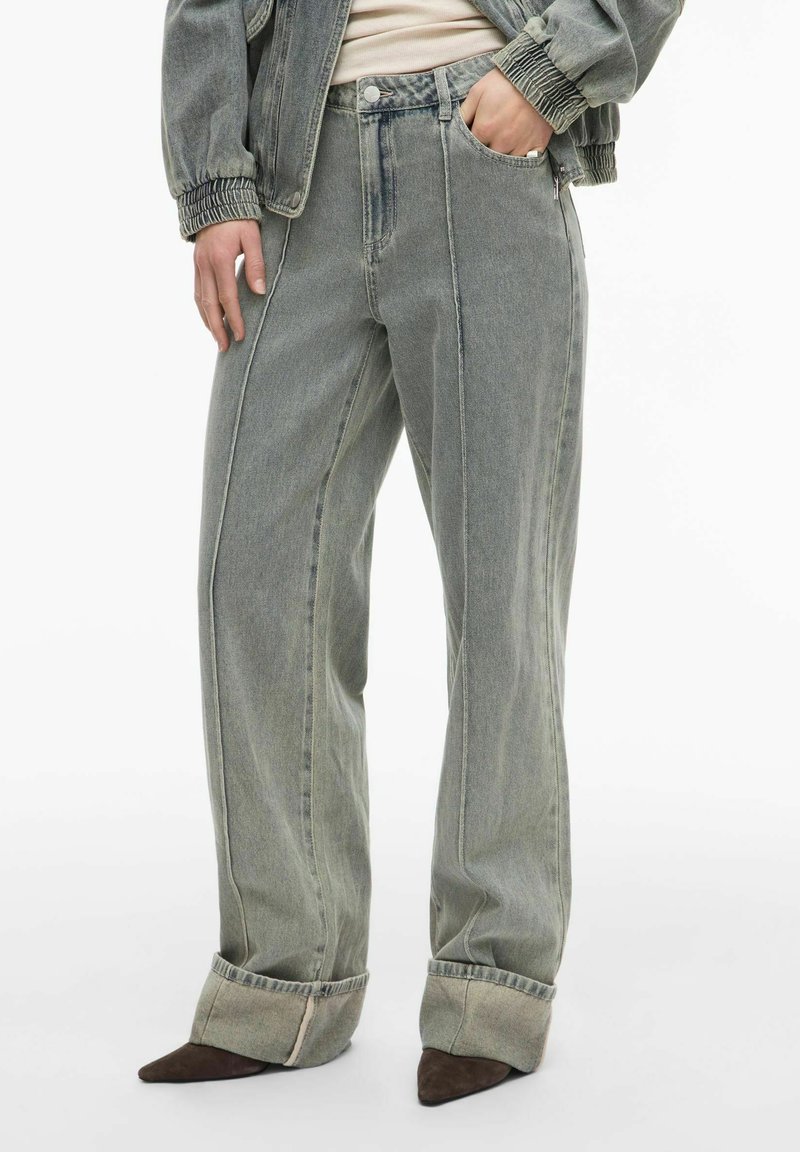 Personne portant un jean gris délavé à taille haute et jambes larges avec ourlets retournés, une veste en jean froncée et des chaussures marron pointues.