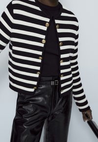 Cardigan a righe bianche e nere con scollo rotondo, dotato di bottoni dorati. Indossato con pantaloni in pelle nera lucida.