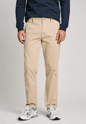 Beige housut, jotka on valmistettu puuvillasta, slim fit -malliset, jossa on nappisulku, vyölenkit ja sivutaskut. Yhdistetty valkoisiin ja harmaisiin lenkkikengiin.